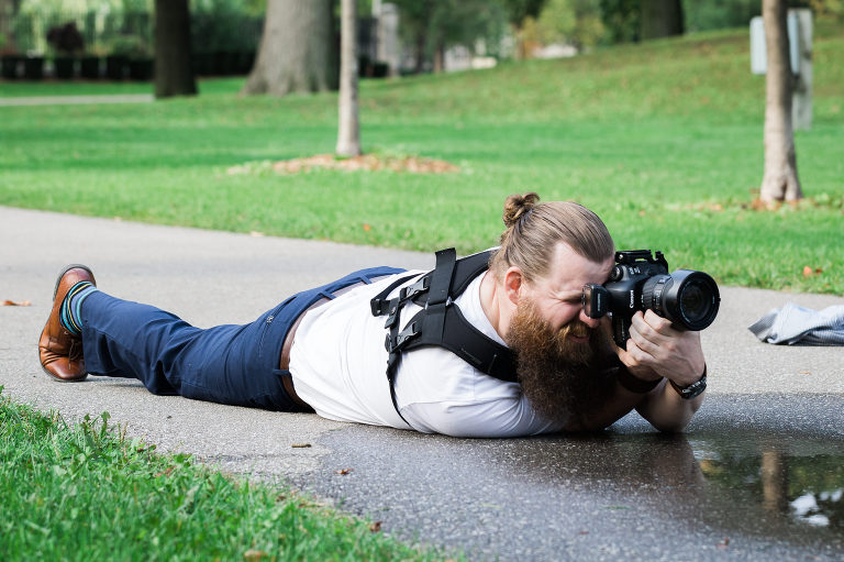 Well dressed male wedding photographer laying on an asphalt path at the edge of a puddle to get the shot. Pony tail and big beard.