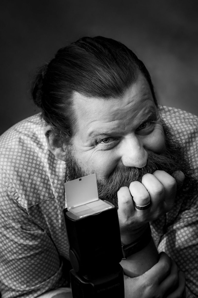 Cheeky black and white photo of male wedding photographer Chad Barry leaning forward with his hair back in a pony tail and other hand on his bearded chin. Twinkle in his eyes.