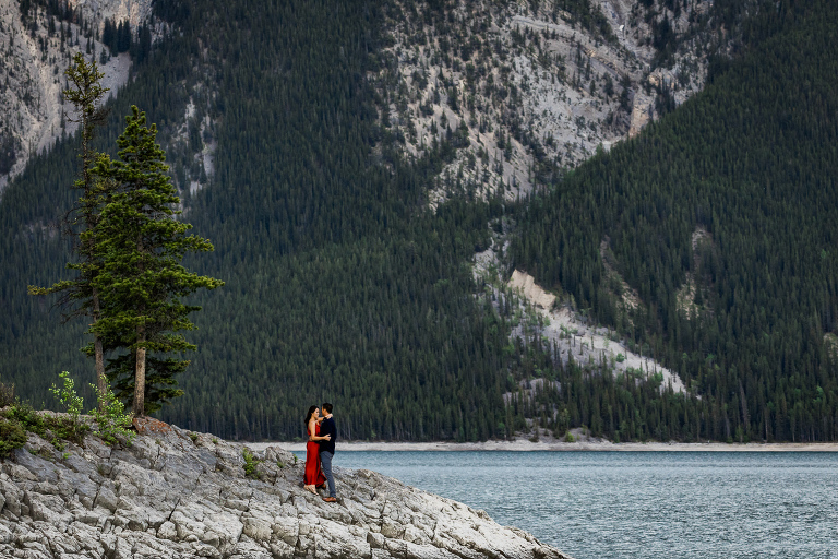Engagements couple embrace on a rocky outcropping with the Rocky Mountains behind them. 
