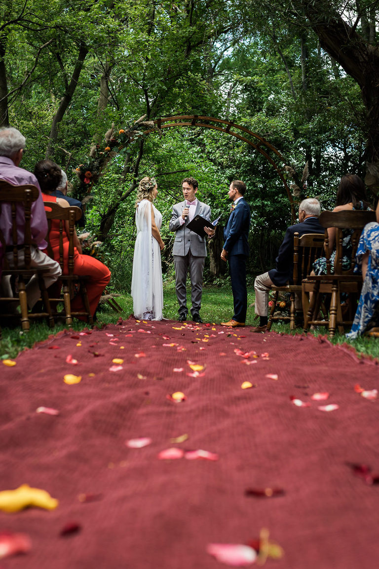 Bride and groom standing at the alter of an outdoor ceremony.  A red carpet lines the aisle covered in flower petals and green trees behind them. 