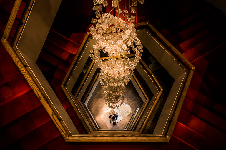 Vintage bride and groom kiss at the bottom of spiral staircase. A giant chandelier hangs down the centre. The staircase is carpeted in deep red. 