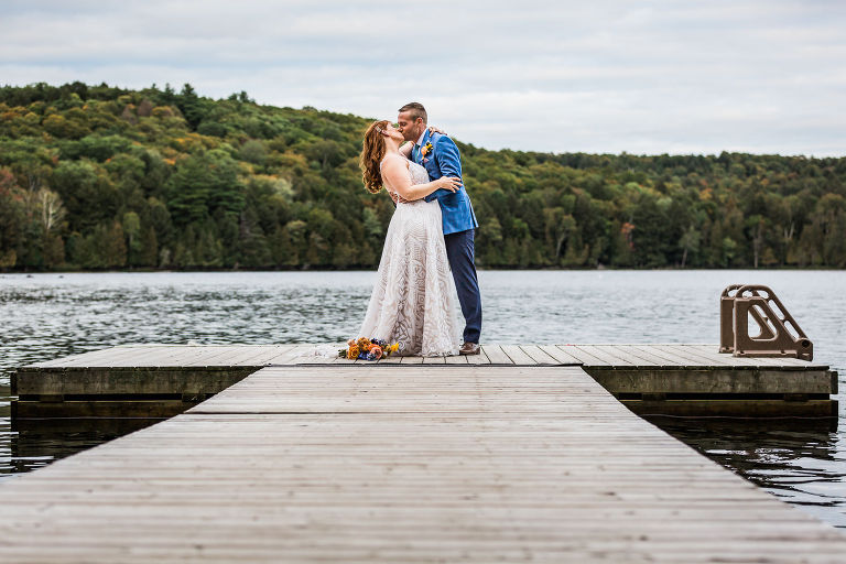 Bride and groom kiss at the end of a dock on a small lake with hills and trees on the far bank. 